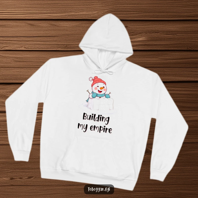 Funny Hoodie: A cheerful snowman comically attempts to build a tiny snowball fort, offering cozy winter humor.